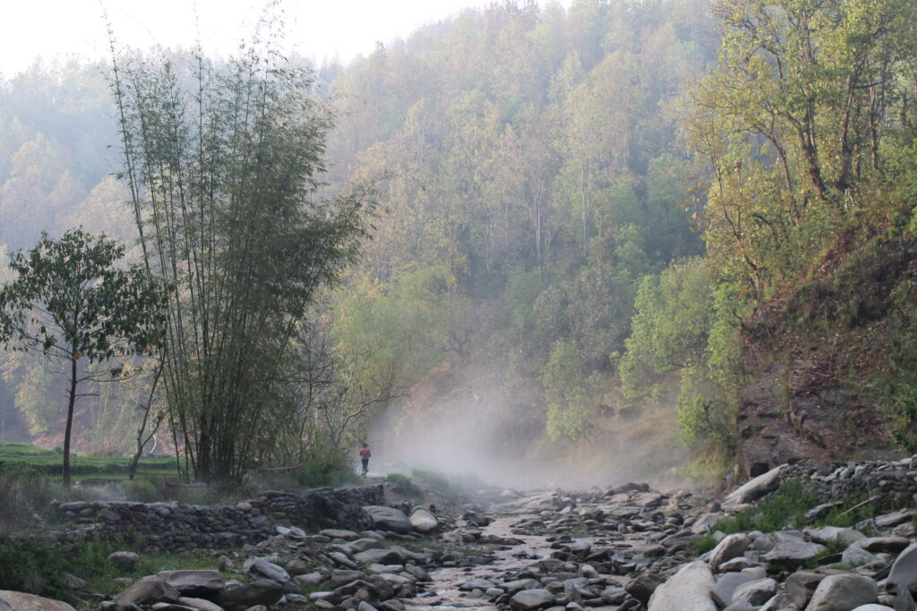 Fog and mist rising over stones in shallow river lined by trees and shrubs.